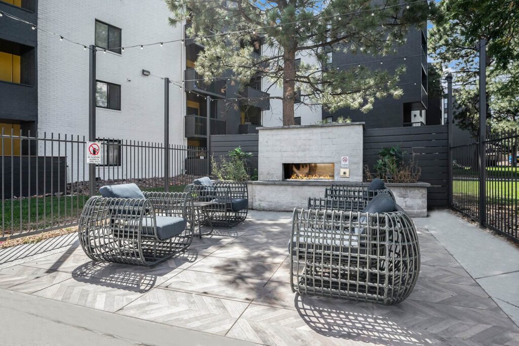 Outdoor seating area with modern metal chairs around a built-in fireplace, surrounded by apartment buildings and greenery.