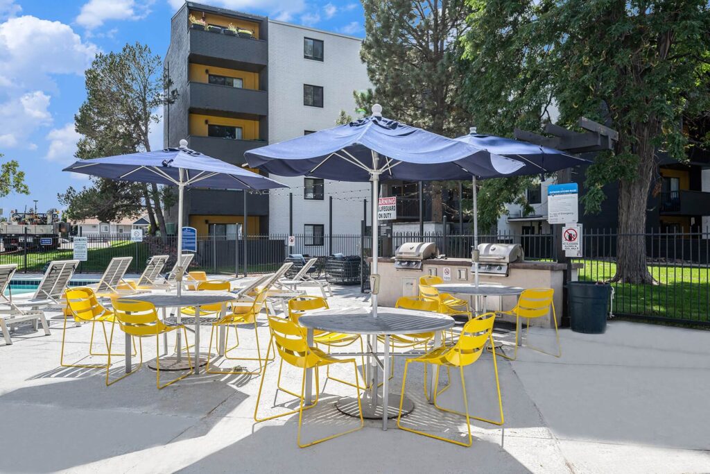 Outdoor apartment patio with blue umbrellas, yellow chairs, and round tables, with nearby barbecue grills and a pool.